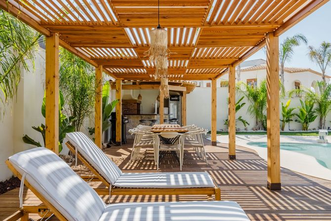 Sunlit patio with wooden pergola, lounge chairs, and a dining table beside a modern house