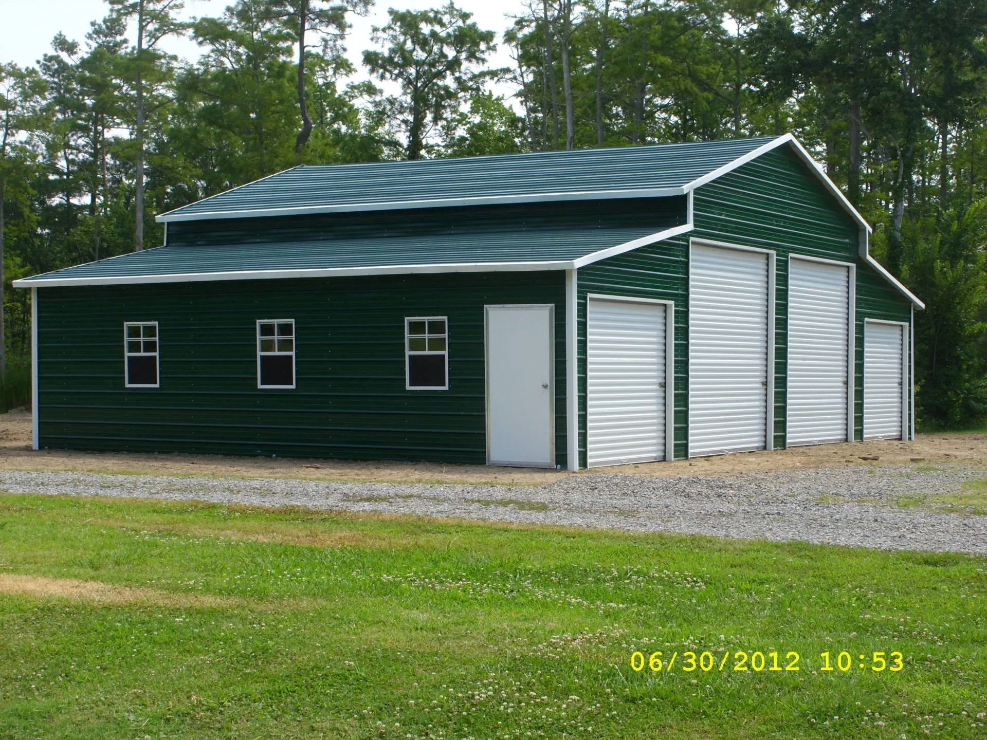 Green metal garage with white doors on a gravel driveway, surrounded by trees.