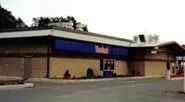 Low brick storefront with blue awning and a parking lot in front