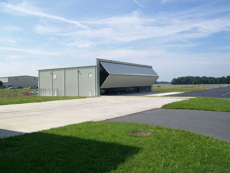 Industrial building with a large gray tilt-up door beside a concrete lot and grass field under a blue sky