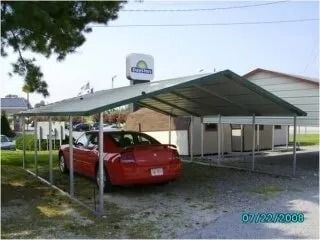 Red car parked under a metal carport beside a small building on a sunny day