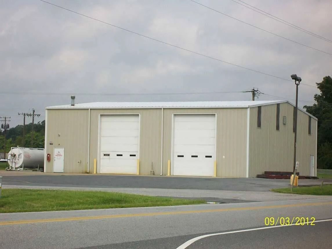 Small beige industrial building with two white garage doors beside a road under cloudy skies.