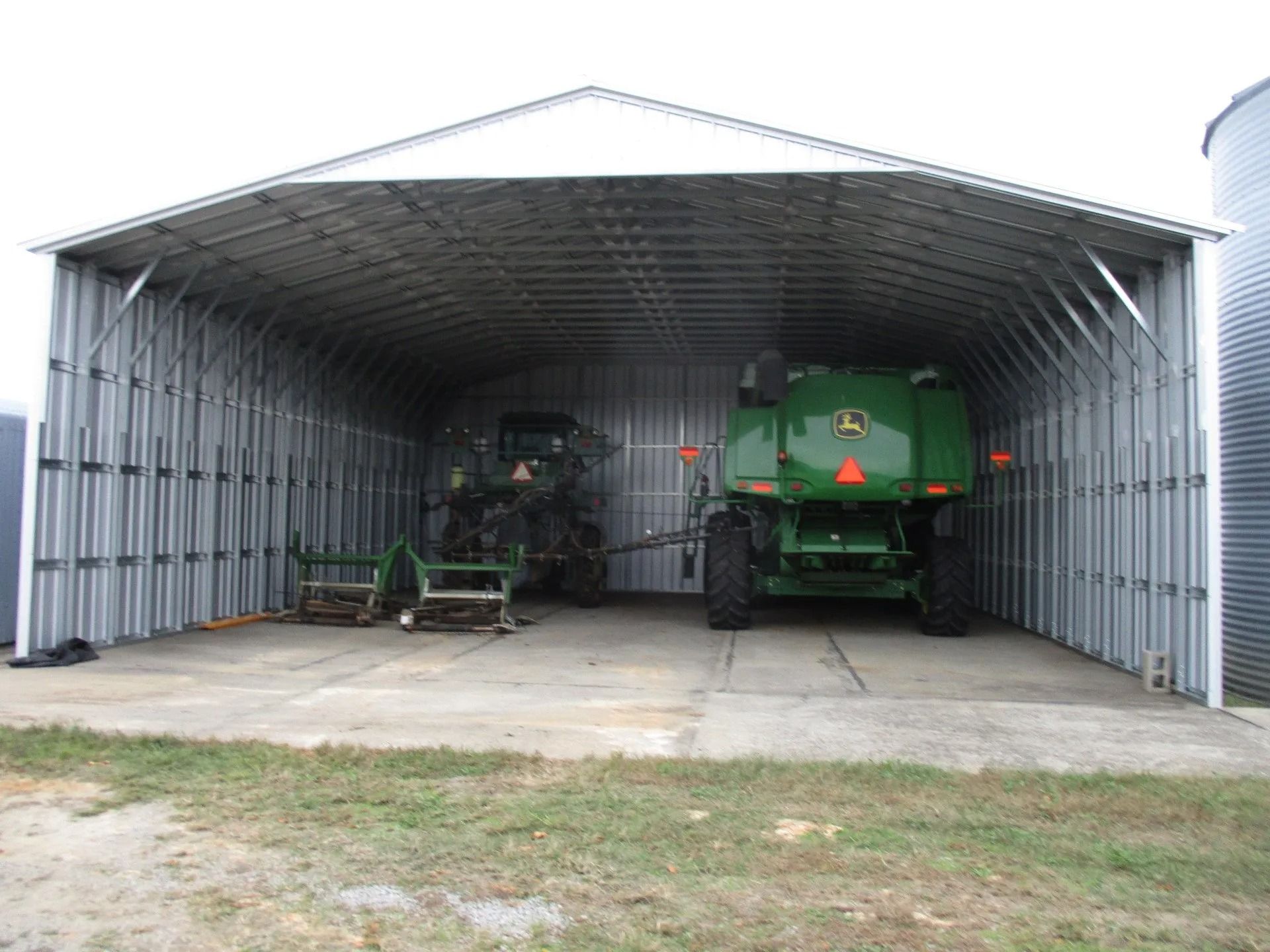Open metal shed with two green farm machines parked inside on a concrete floor.