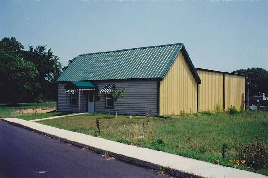 Small yellow-and-gray building with a green roof beside a road and grassy lot.