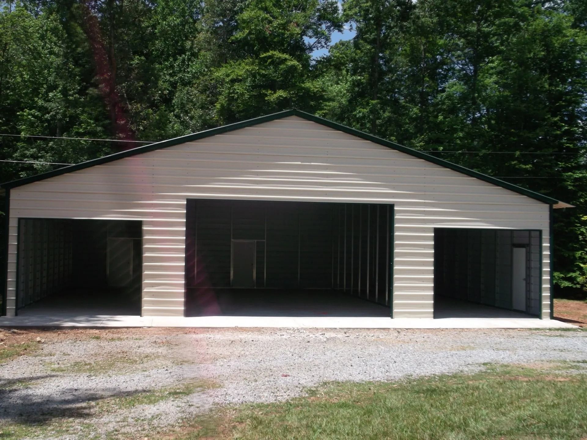 White garage with three open bays and dark interiors, set beside trees and a gravel driveway.