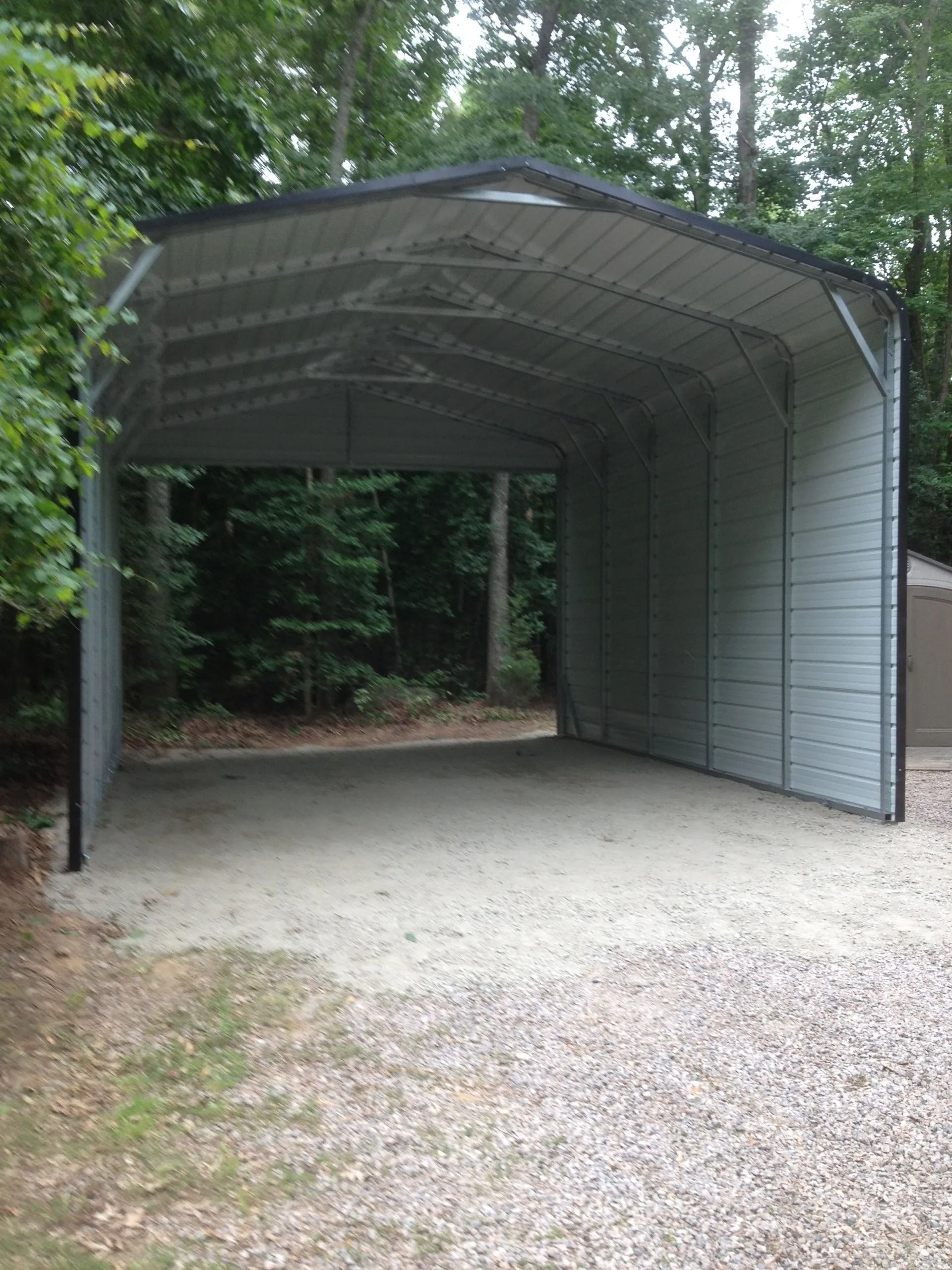 Metal carport or covered parking shelter with gravel floor at a wooded driveway entrance