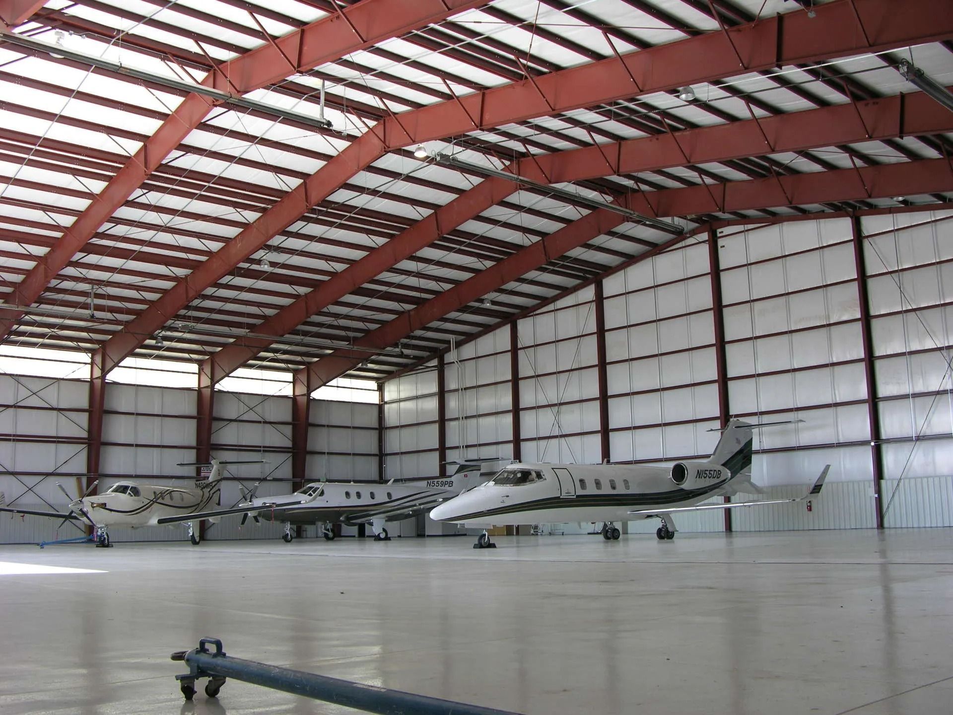 Small aircraft parked inside a large hangar with a red arched roof and open concrete floor.
