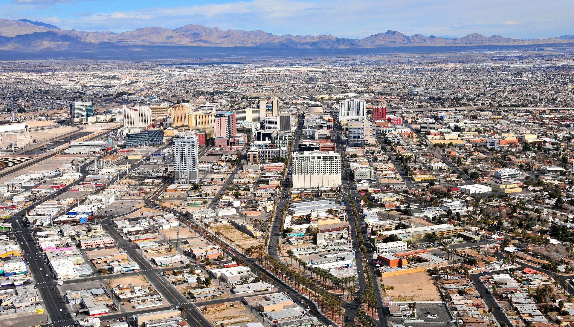 Aerial view of downtown Las Vegas with tall buildings and a long, straight road, with mountains in the background.