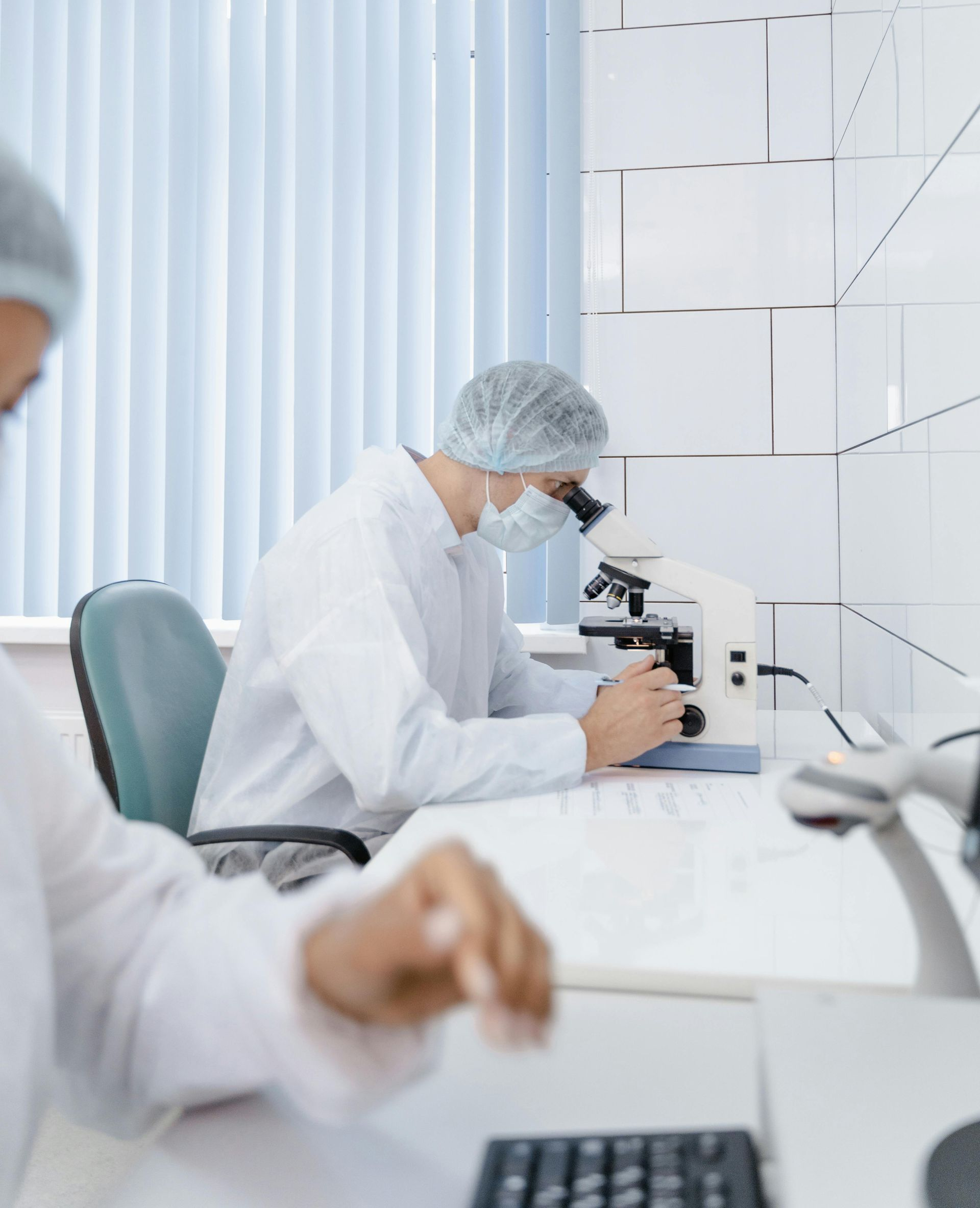Scientist in lab coat and hairnet looks into a microscope evaluating a mold sample