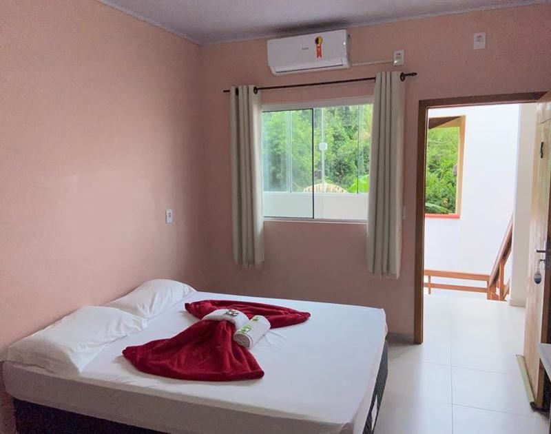 Um quarto com paredes rosa-claro, uma cama branca com uma toalha vermelha como decoração, uma janela com cortinas e uma porta aberta.