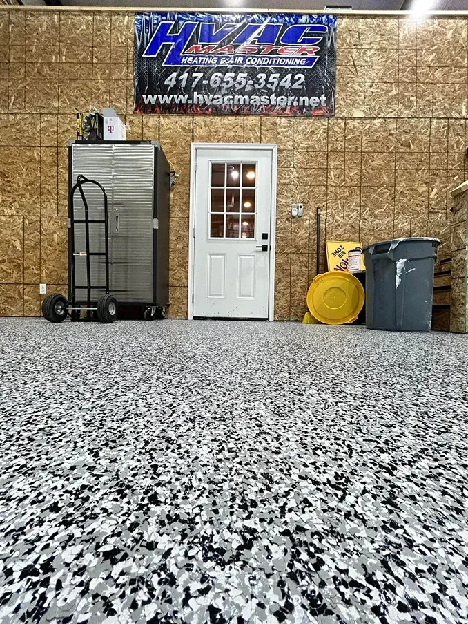 Garage interior with white door, large banner, hand truck, barrels, and speckled epoxy floor.