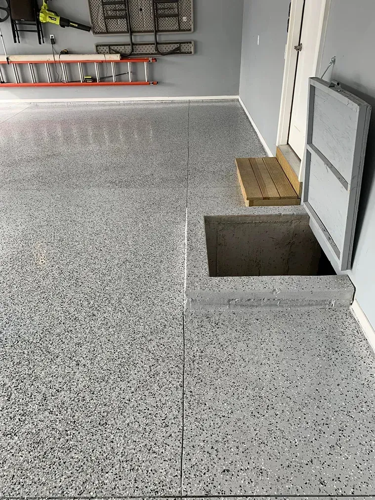 Speckled garage floor with an open square floor hatch near a wall and step.