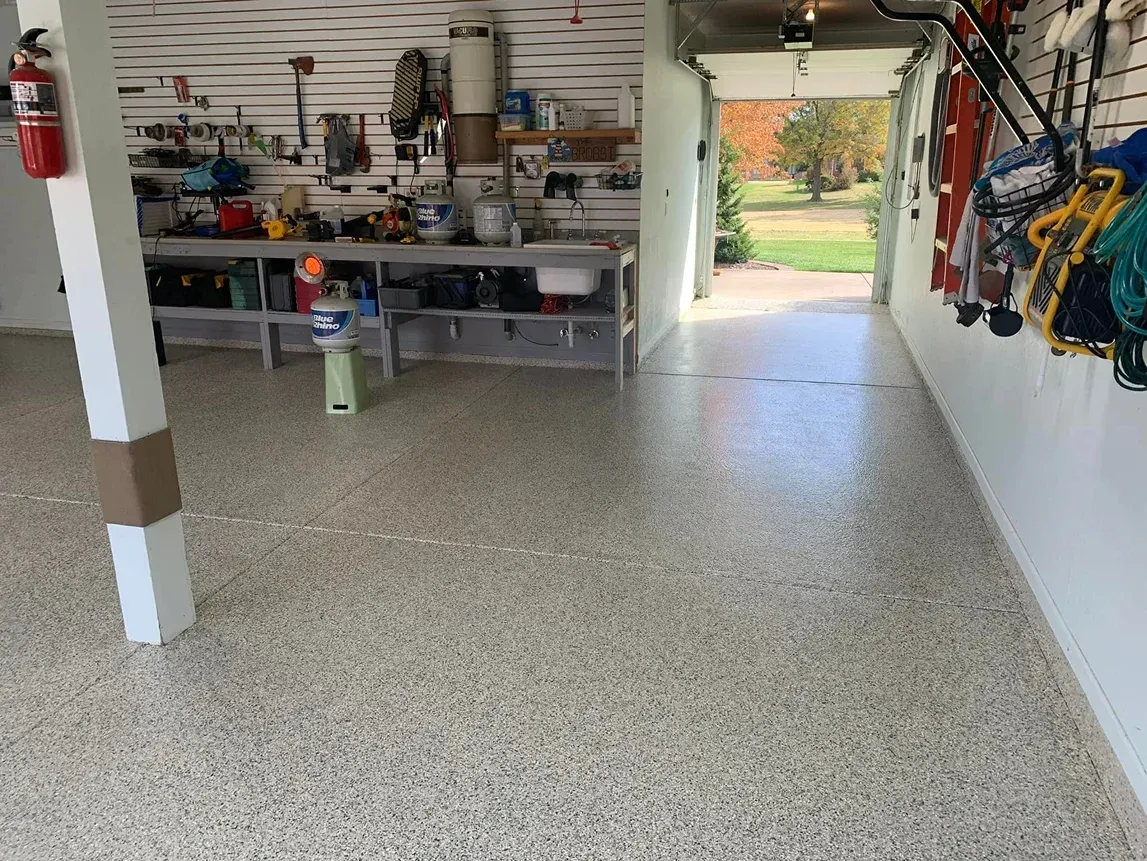 Garage with speckled epoxy floor, shelves of tools and supplies, and hanging sports gear near an open doorway.
