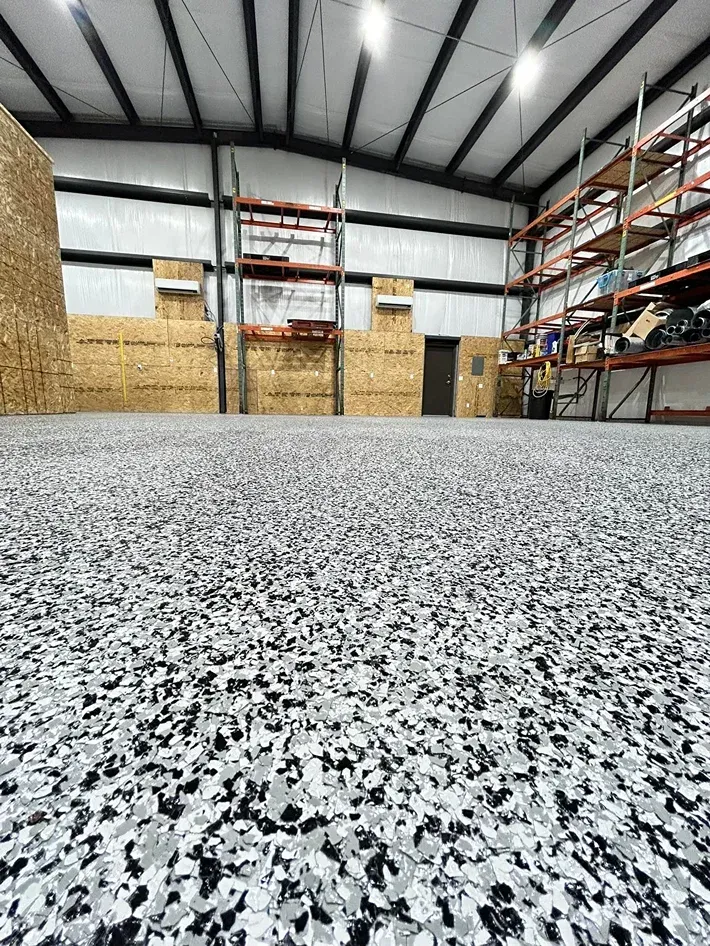 Empty warehouse with speckled epoxy floor, metal shelving, and stacked cardboard boxes under high ceiling lights