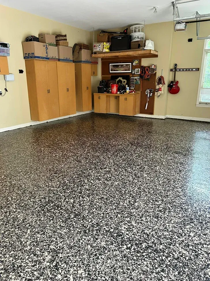 Empty garage with speckled epoxy floor, wooden cabinets, shelves, and hanging tools.
