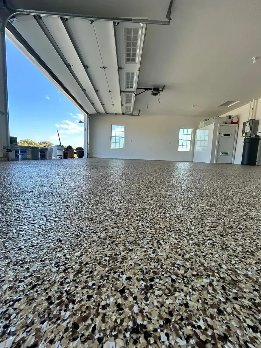 Empty garage with speckled epoxy floor, white walls, open ceiling beams, and bright windows.