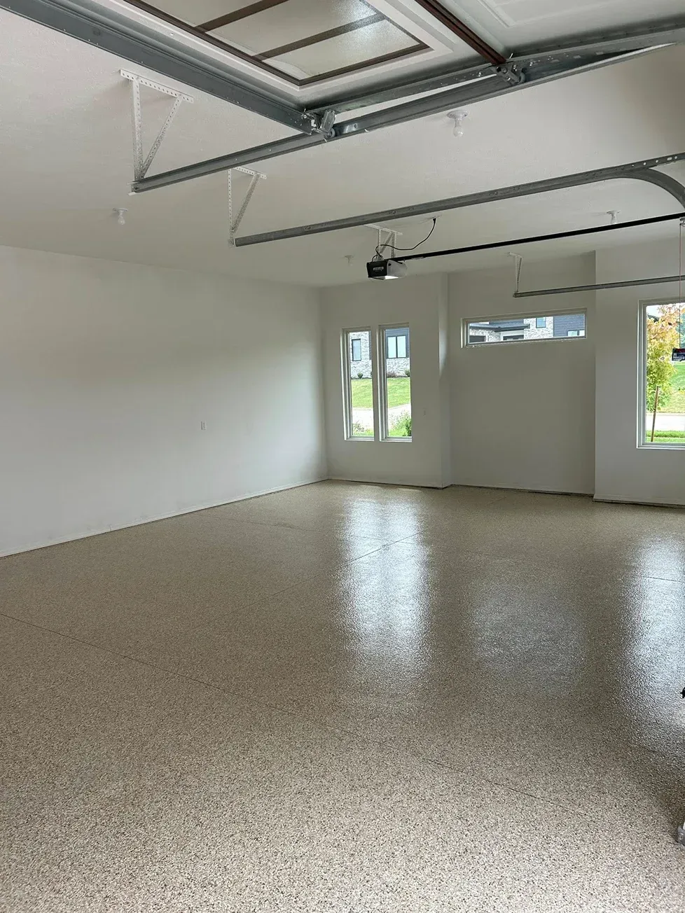 Empty garage interior with white walls, speckled floor, exposed ceiling beams, and windows on the far wall