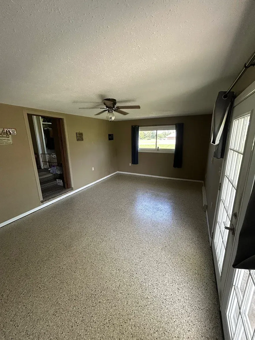 Empty living room with beige walls, speckled floor, ceiling fan, and windows letting in daylight