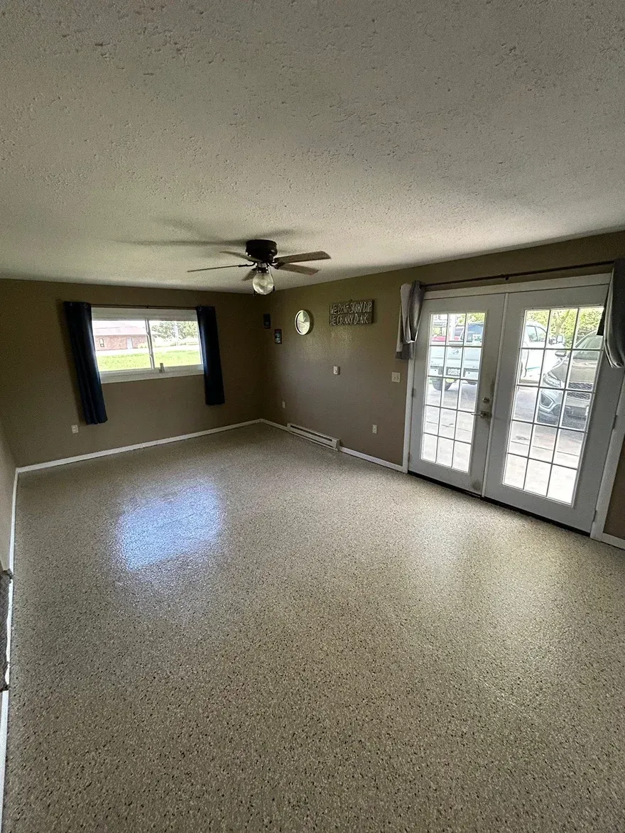 Empty living room with speckled floor, ceiling fan, window, and glass double doors.