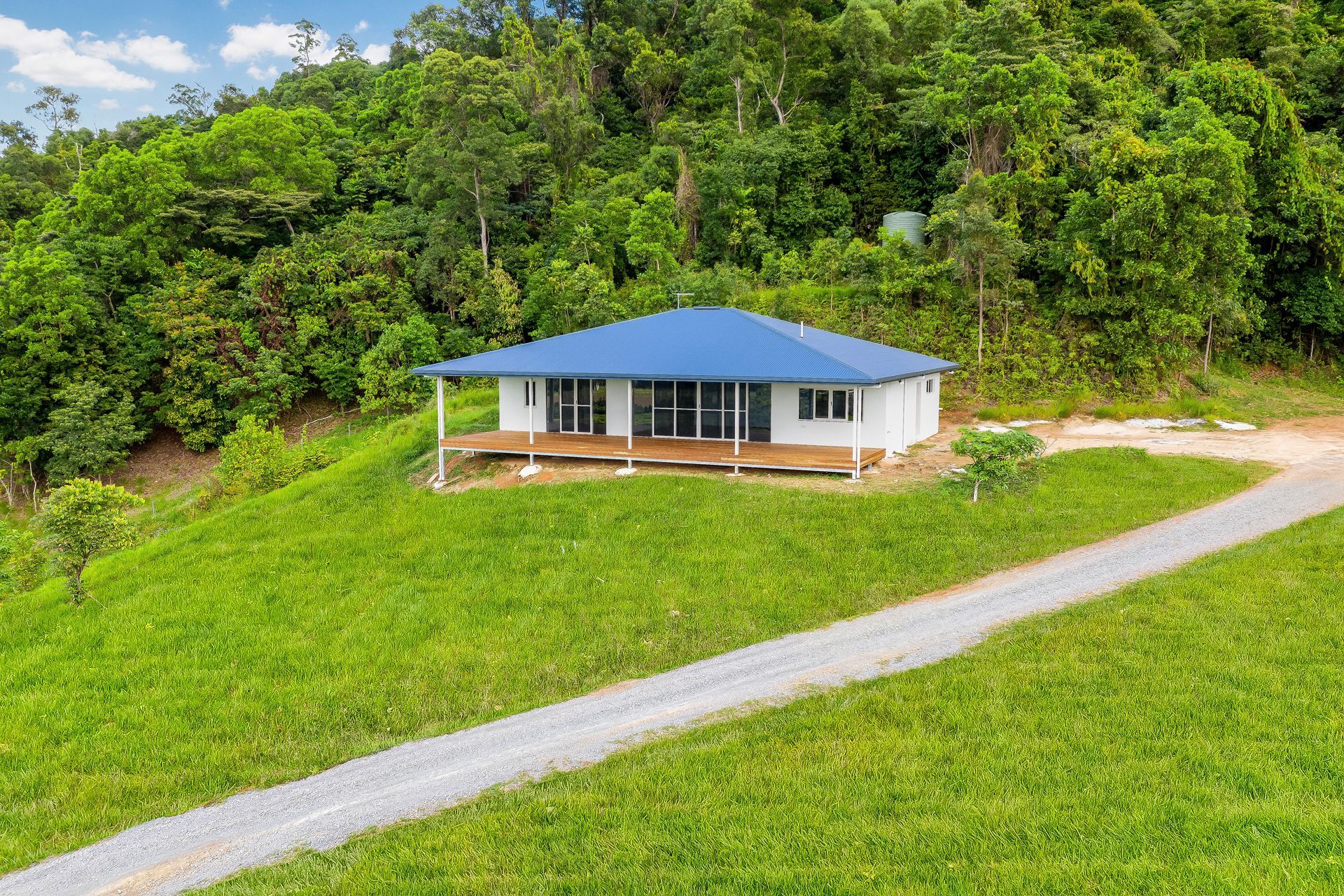 A White House With a Blue Roof Sits on a Grassy Hill in Front of a Lush — Ashlee Jones Homes in Gordonvale, QLD