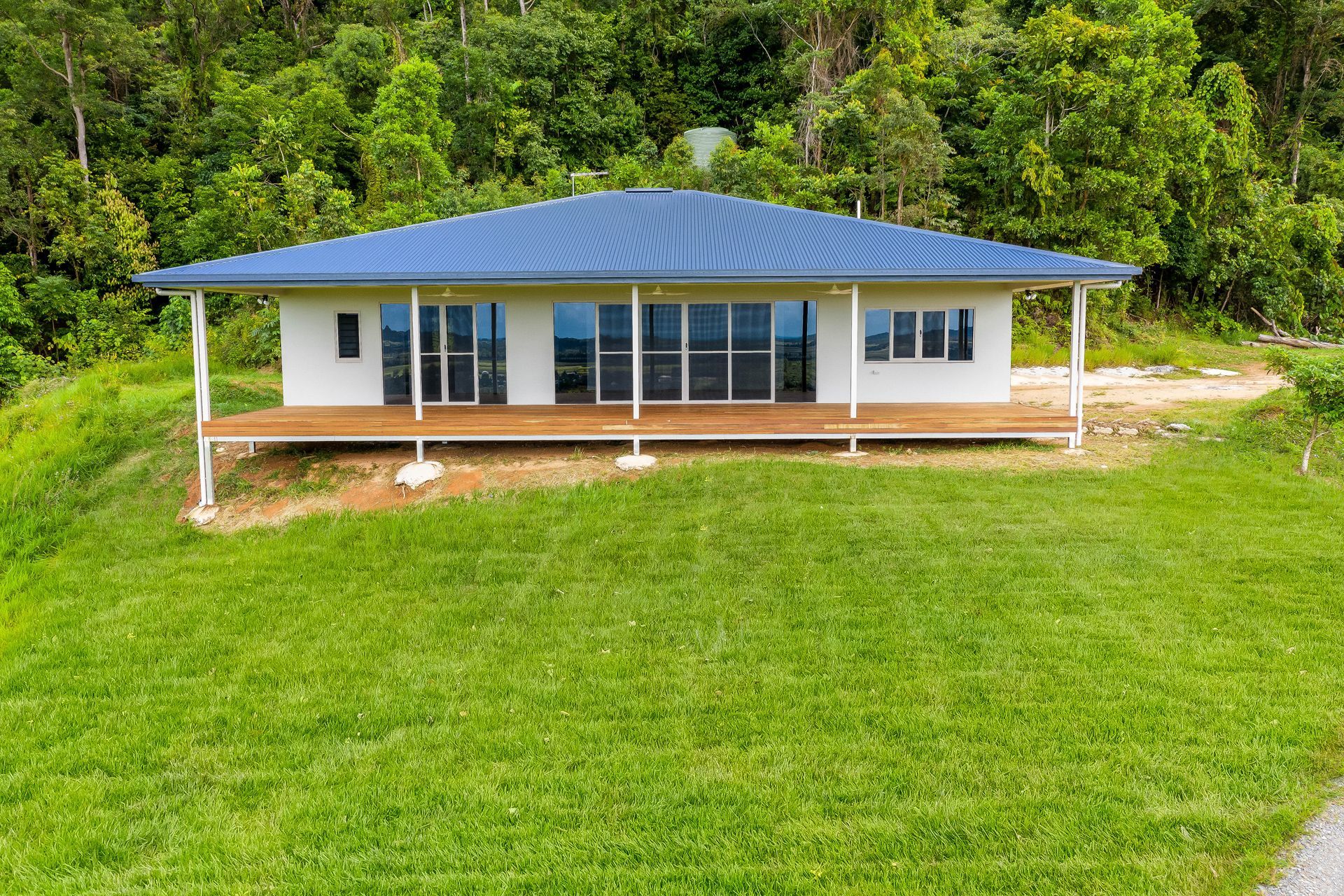 A White House With a Blue Hip Roof and a Spacious Wooden Deck Sits — Ashlee Jones Homes in Gordonvale, QLD