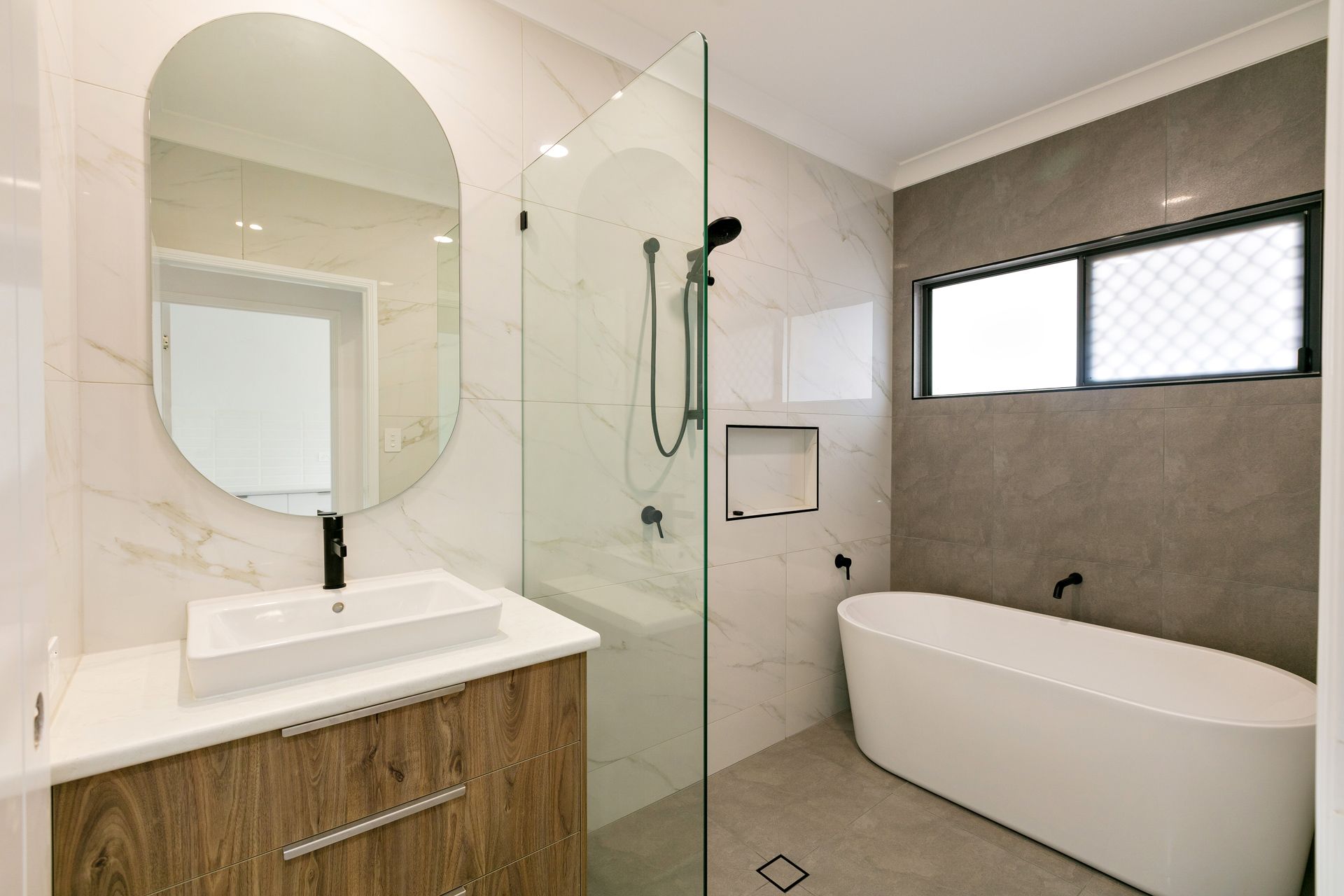 A Modern Bathroom With a Wood Vanity, an Oval Mirror — Ashlee Jones Homes in Gordonvale, QLD