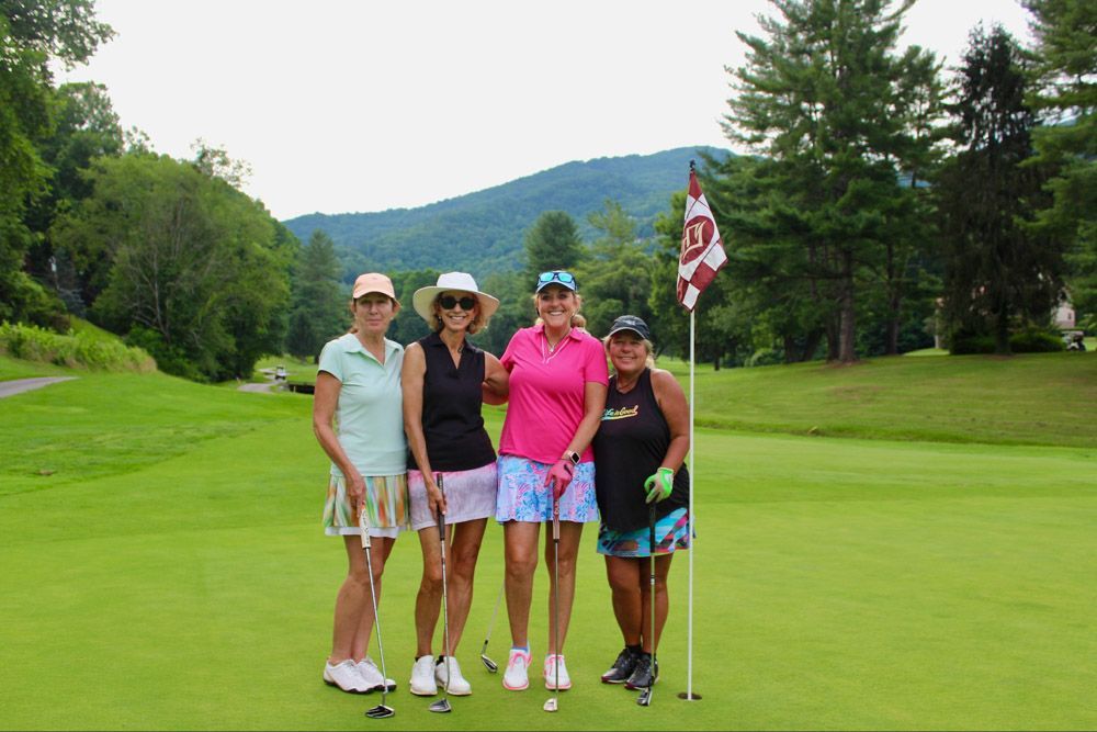 Four golfers stand together on a putting green near a flag, with a mountain and trees in the background.