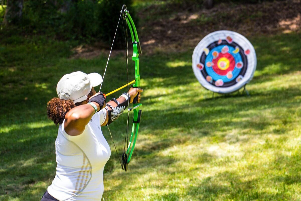 Archer in a white shirt and cap aiming a bright green bow at a target outdoors