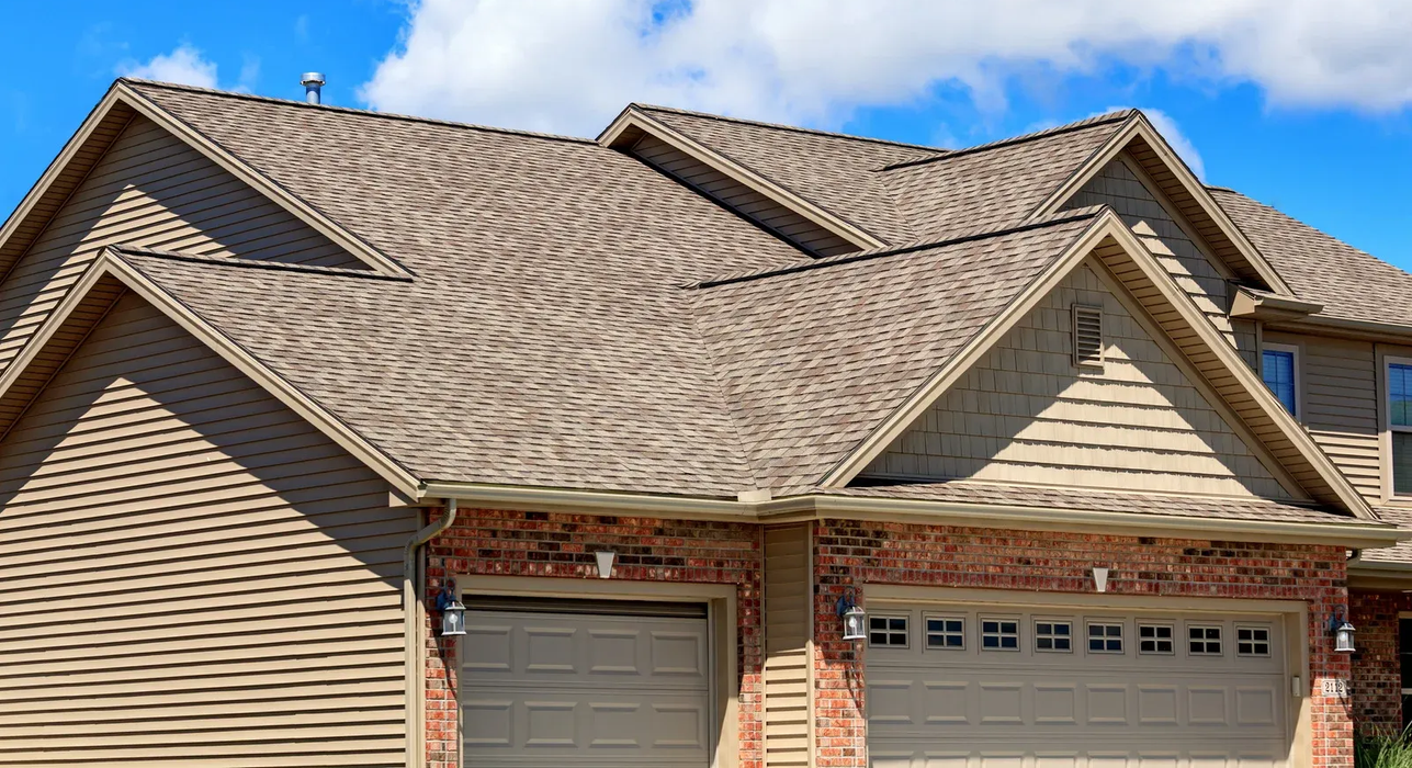 Tan shingle roof on a house with beige siding, brick facade, and two garage doors; bright blue sky with clouds.