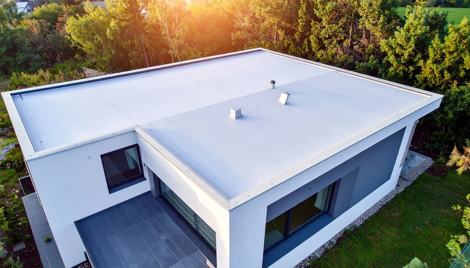 Modern house with a flat white roof, gray siding, and a wooden deck, surrounded by trees.