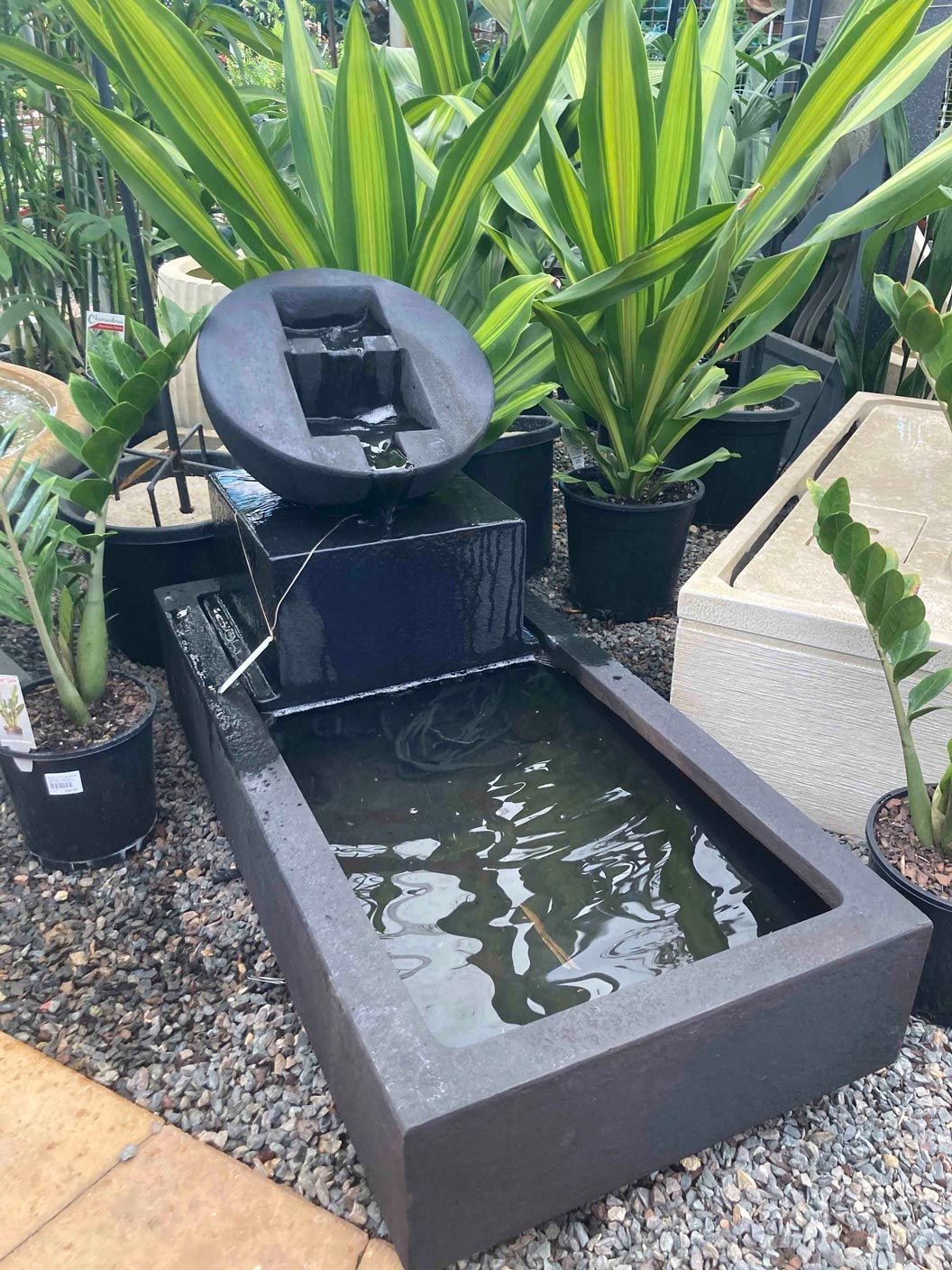 Small outdoor water fountain with dark stone basin, spouting water, surrounded by lush green plants.