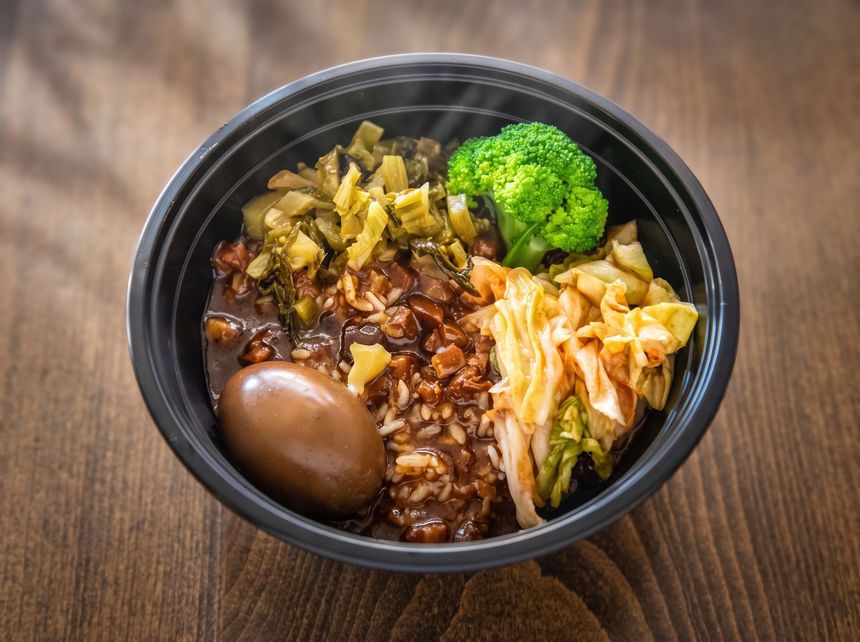 Black bowl of rice with vegetables, broccoli, dumpling, and a brown egg on a wooden table