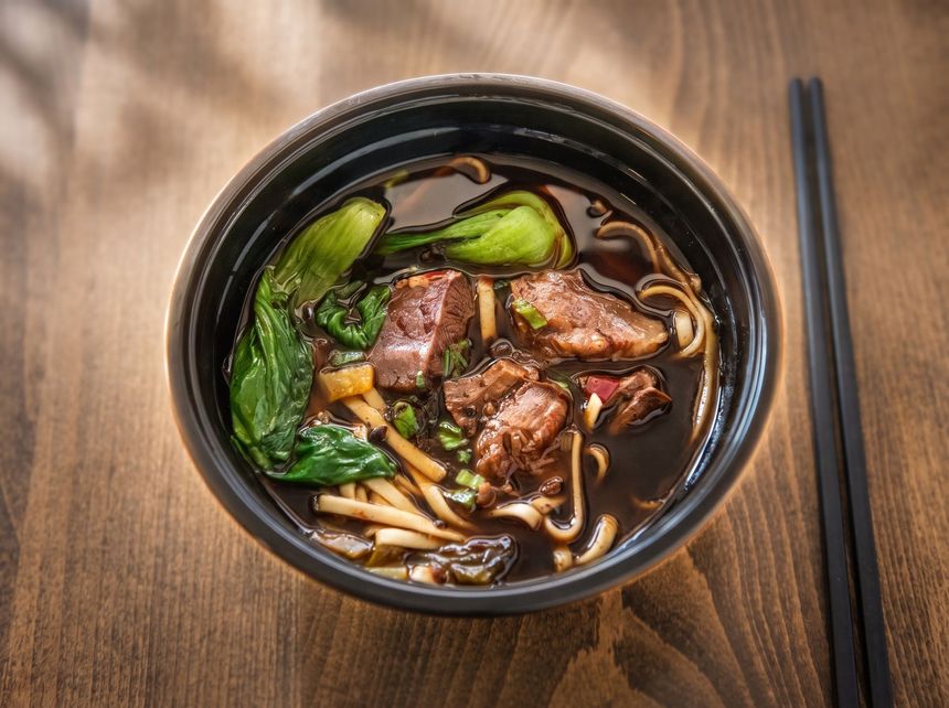 Black bowl of beef noodle soup with greens on a wooden table, chopsticks beside it