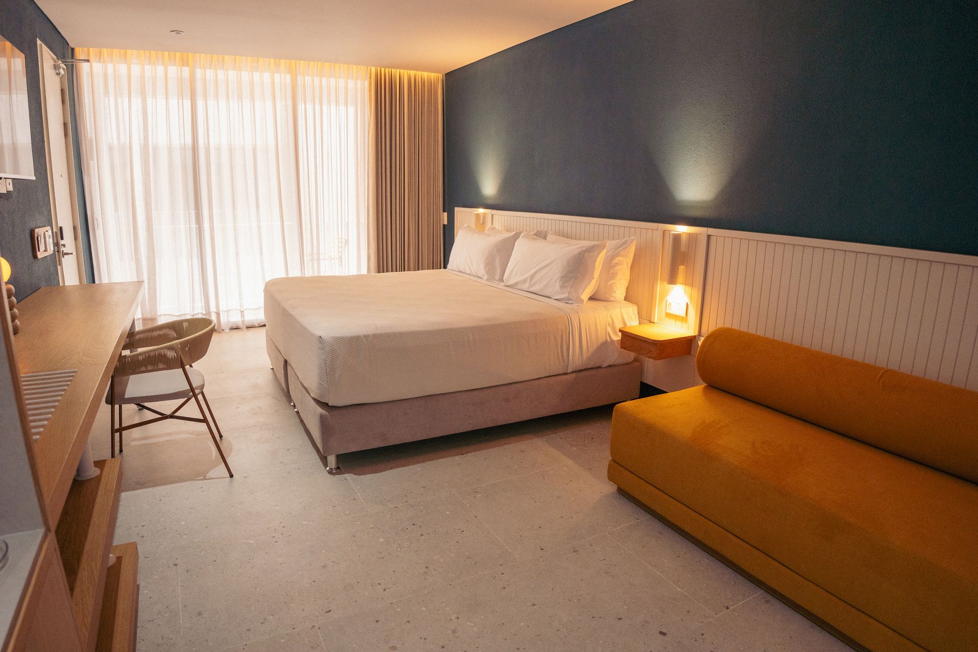 habitaciones en san andres, con cama amplia, sofá amplio color mostaza, cortinas claras y pared azul en ambiente moderno.