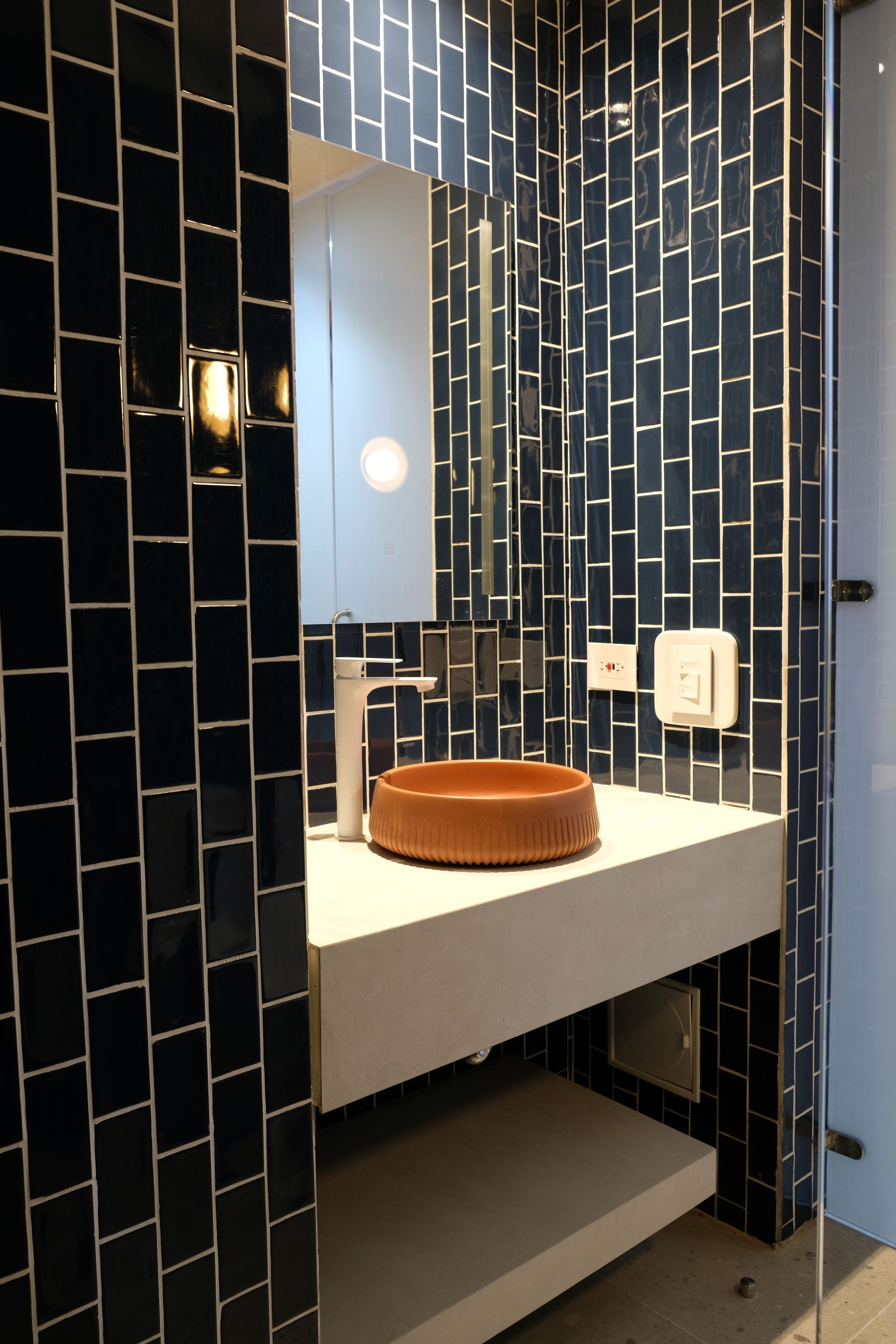 Baño con lavamanos redondo color terracota, grifería blanca, espejo y azulejos azul oscuro.