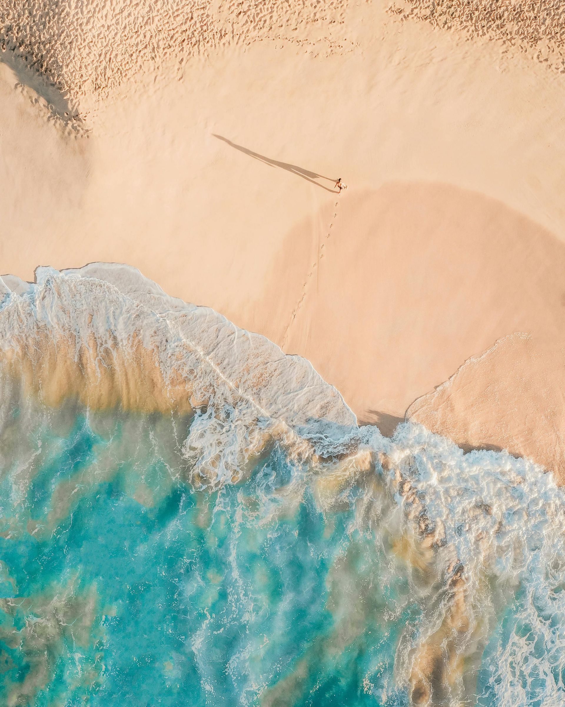 Vista aérea de praia com areia clara, mar tranquilo e ondas suaves tocando a costa em ambiente tropical