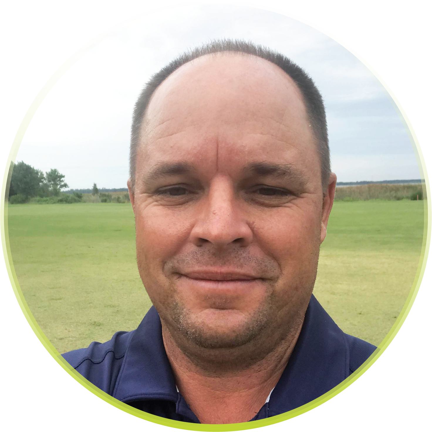 A man in a blue shirt is smiling in front of a green field.