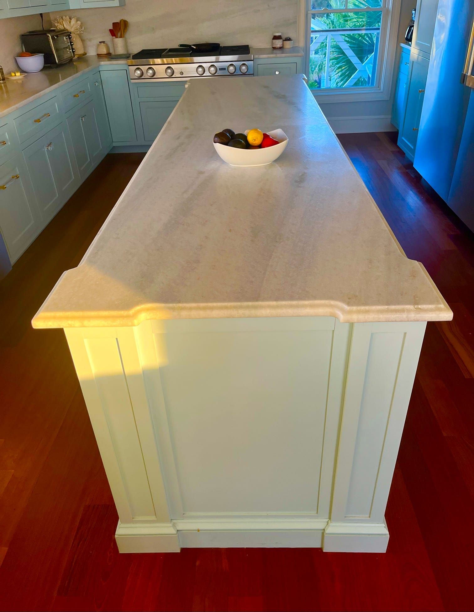 Bright kitchen with a large white island and bowl of fruit on top, light blue cabinets, and wood floors.