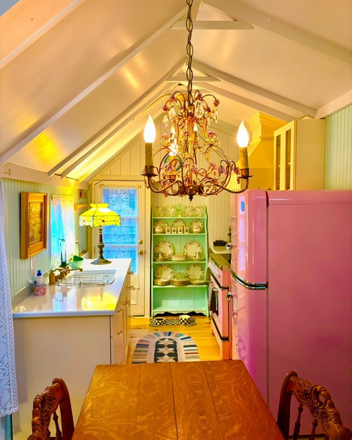 Bright, narrow kitchen with pink fridge, green shelves, white counters, and a chandelier over a wooden table