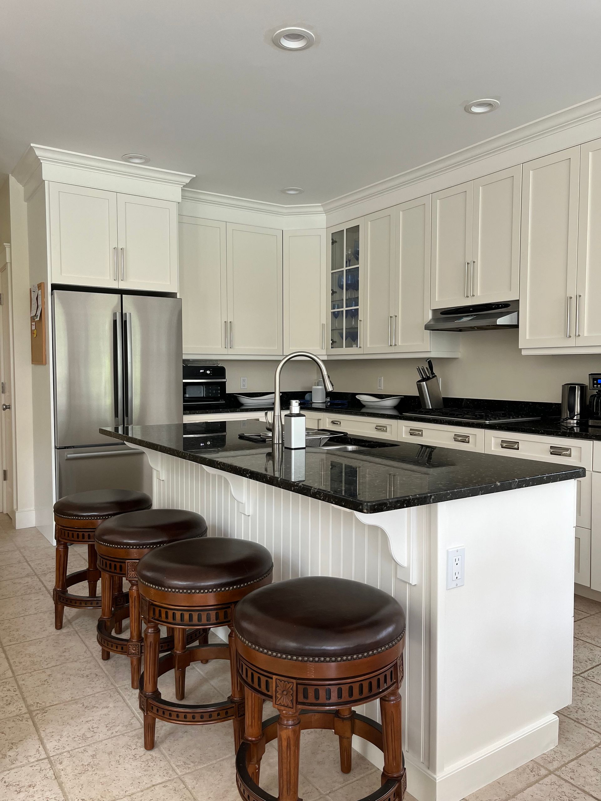 Bright white kitchen with black granite island, stainless fridge, and three wooden stools