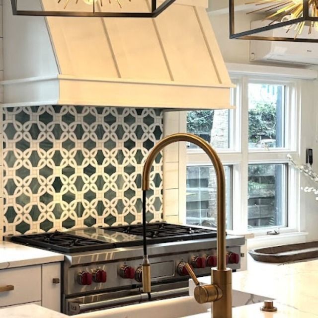 Modern kitchen with patterned tile backsplash, stainless range hood, and brass faucet at island sink