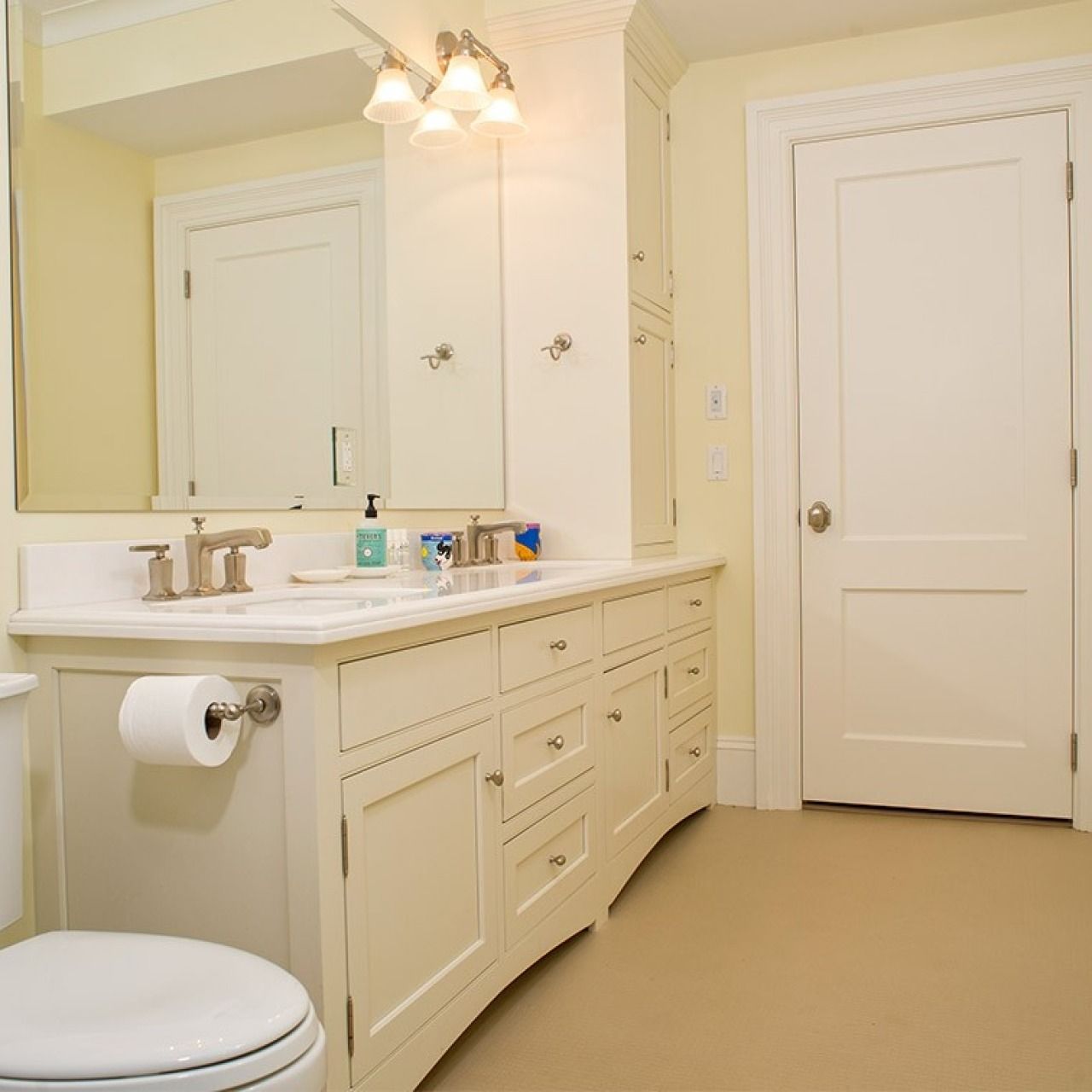 Bright bathroom with white vanity, large mirror, sink, toilet paper holder, and closed door on beige floor.