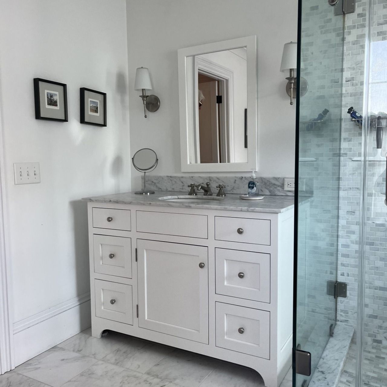 Bright white bathroom vanity with mirror, marble countertop, sconces, and glass shower enclosure