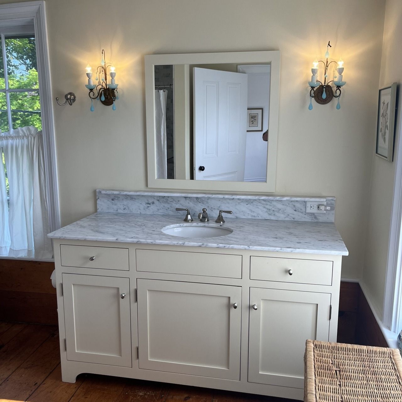 White bathroom vanity with marble top, mirror, wall sconces, and a woven basket in a bright room