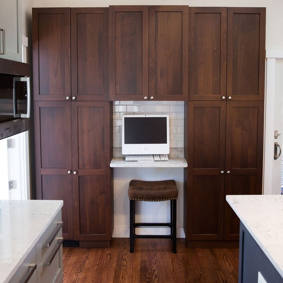 Kitchen wall of dark wood cabinets with a small built-in desk, monitor, and stool in the center