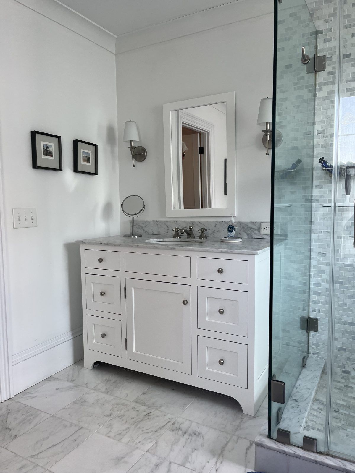 Bright white bathroom vanity with marble countertop, mirror, wall sconces, and glass shower door