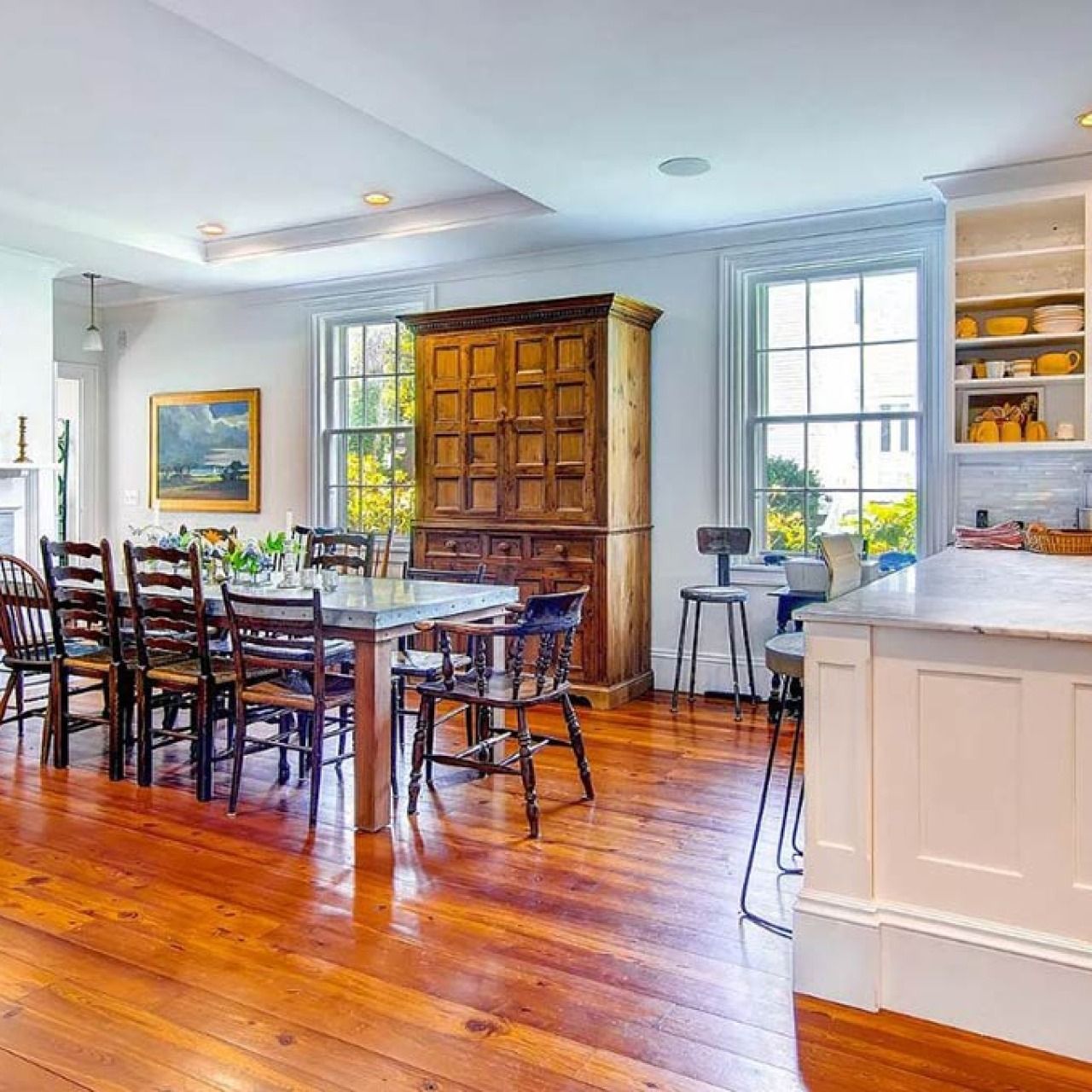 Bright dining room with wooden table, chairs, armoire, and polished hardwood floors near large windows.