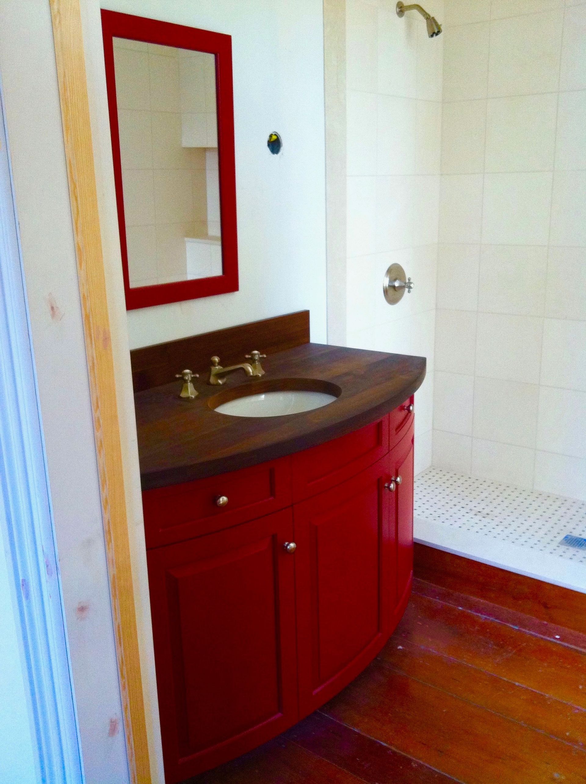 Small bathroom with red vanity, mirror, sink, and a bathtub on a wooden floor.