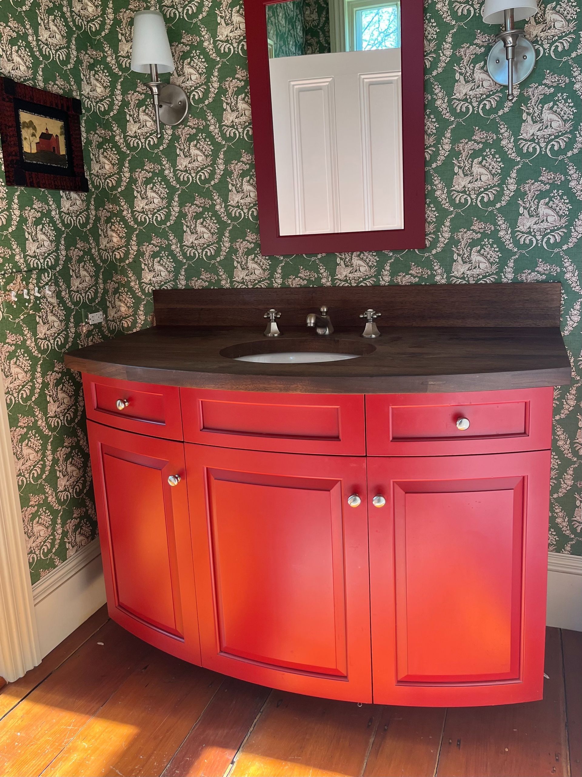Red vanity sink with mirror in a wallpapered bathroom, lit by sunlight from the left.