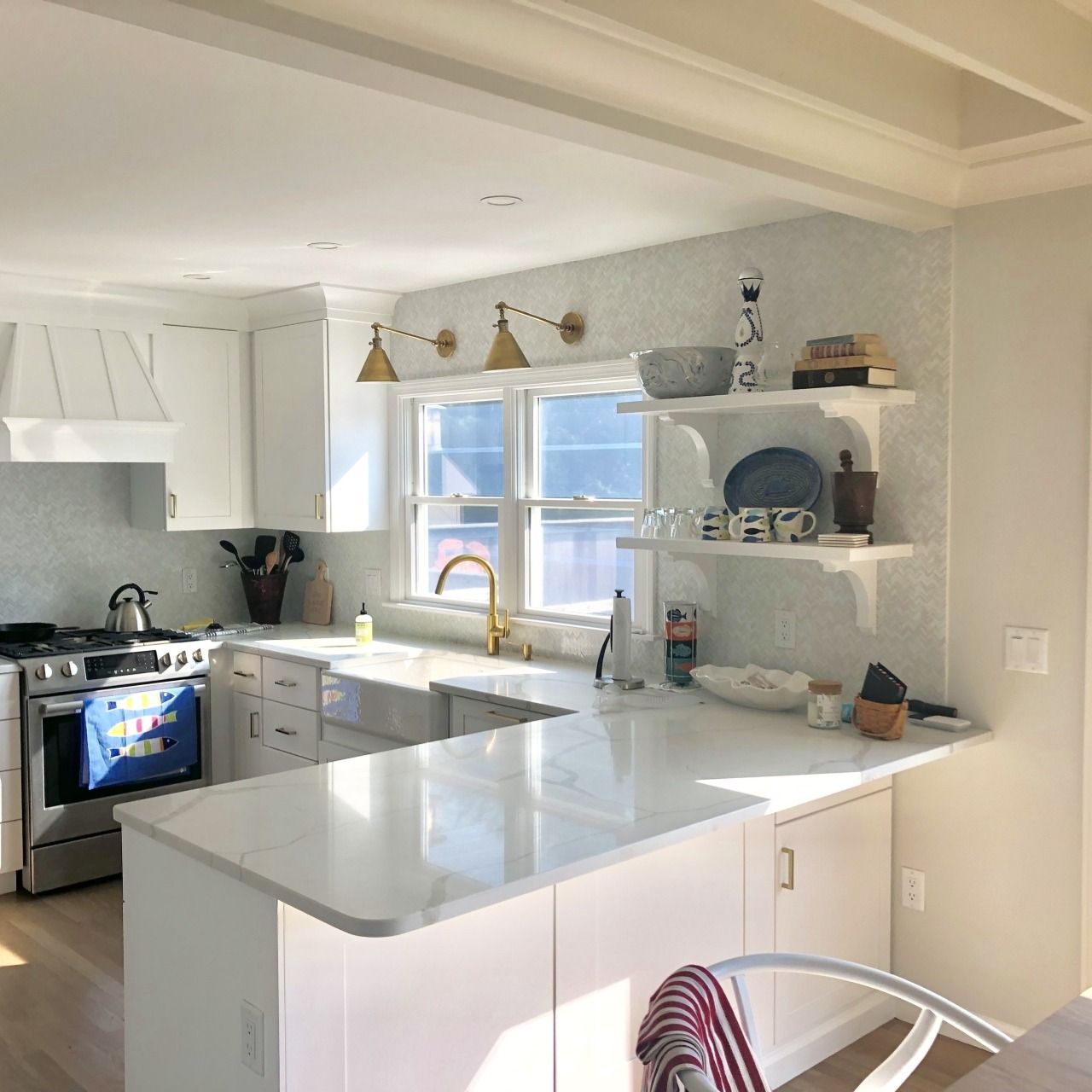 Bright white kitchen with island, gold faucet, open shelves, and sunlight through a window.